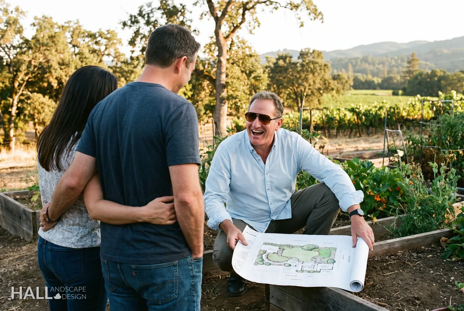 Ian Hall crouched at a garden bed with the landscape plan unrolled, laughing with a couple during their on-site consultation