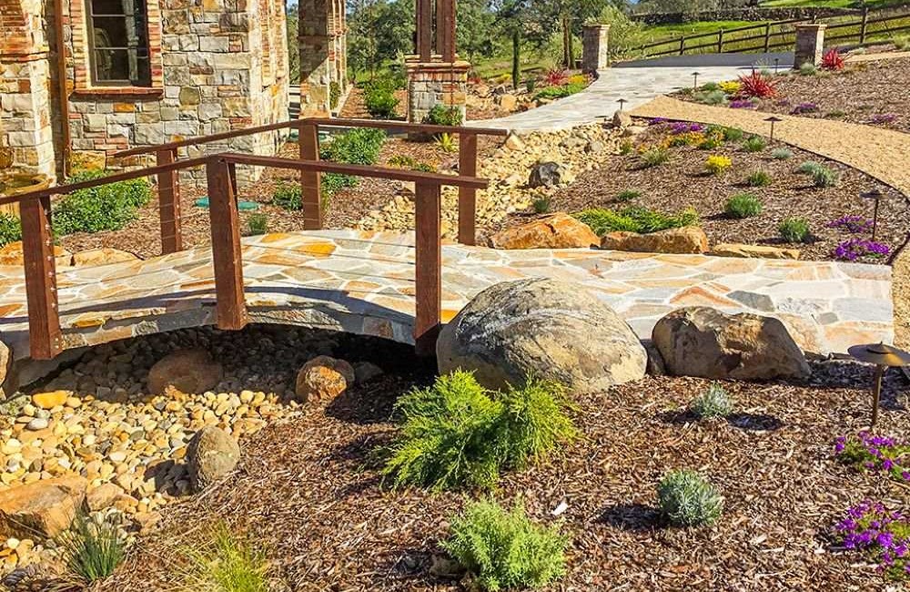 xeriscape yard with colorful plants and brick footbridge over dry stream bed. 