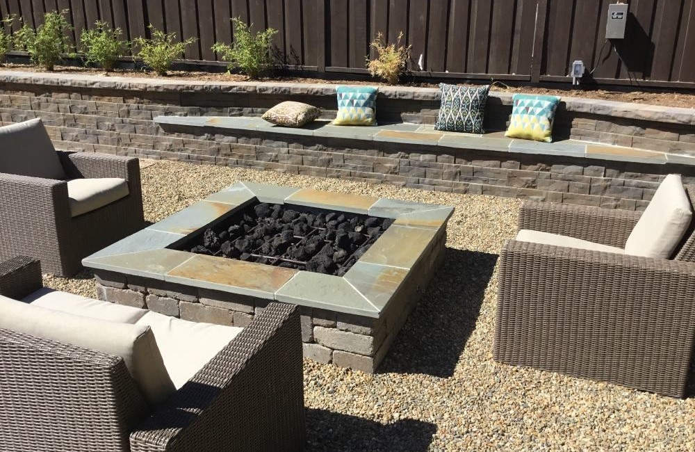 Square fire pit of brick with custom cement top and black "coals." Three chairs and cushions on retaining wall. 