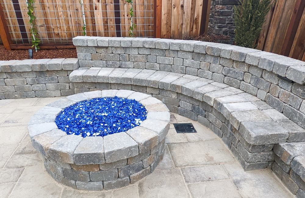 Round brick fire pit with blue "coals" surrounded on one side with retaining wall bench. 