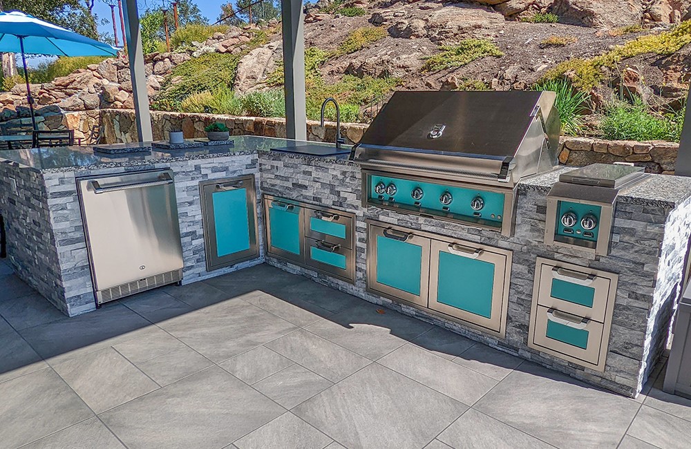 Grey  multi-colored brick outdoor kitchen with turquoise accents on grey patio with xeriscape above retaining wall behind. 