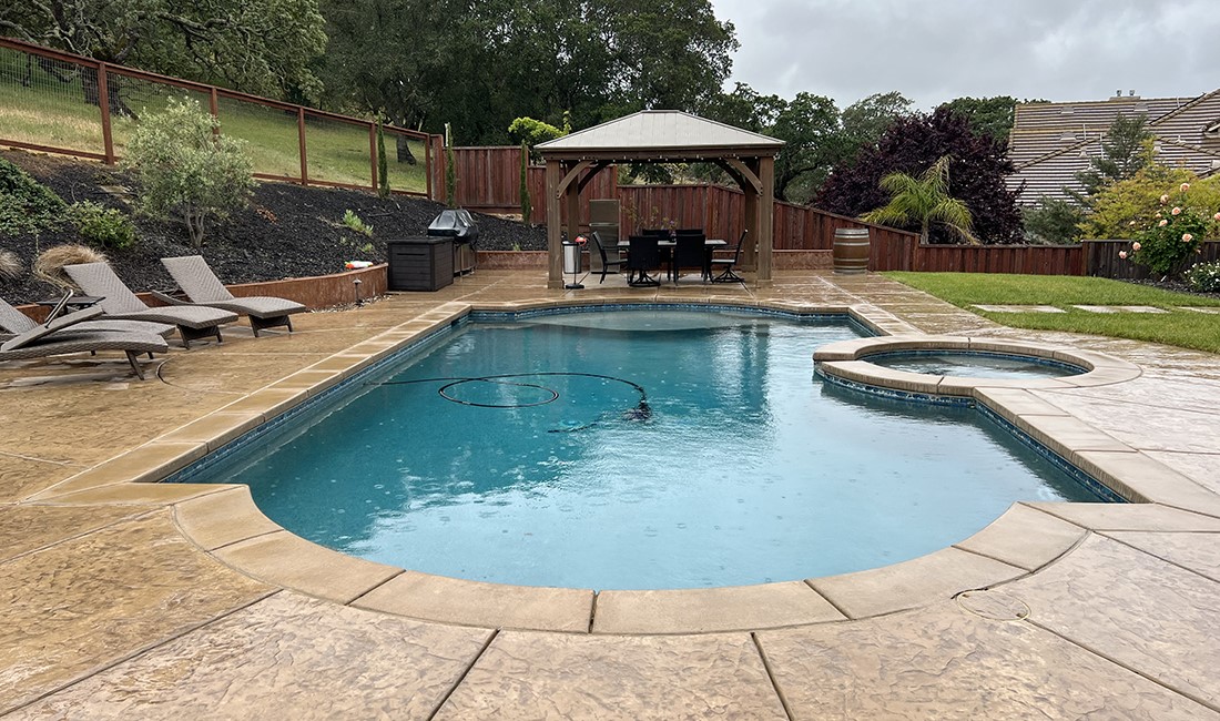 Custom-shape pool with inset spa surrounded by hardscape pool deck, in front of pergola. 