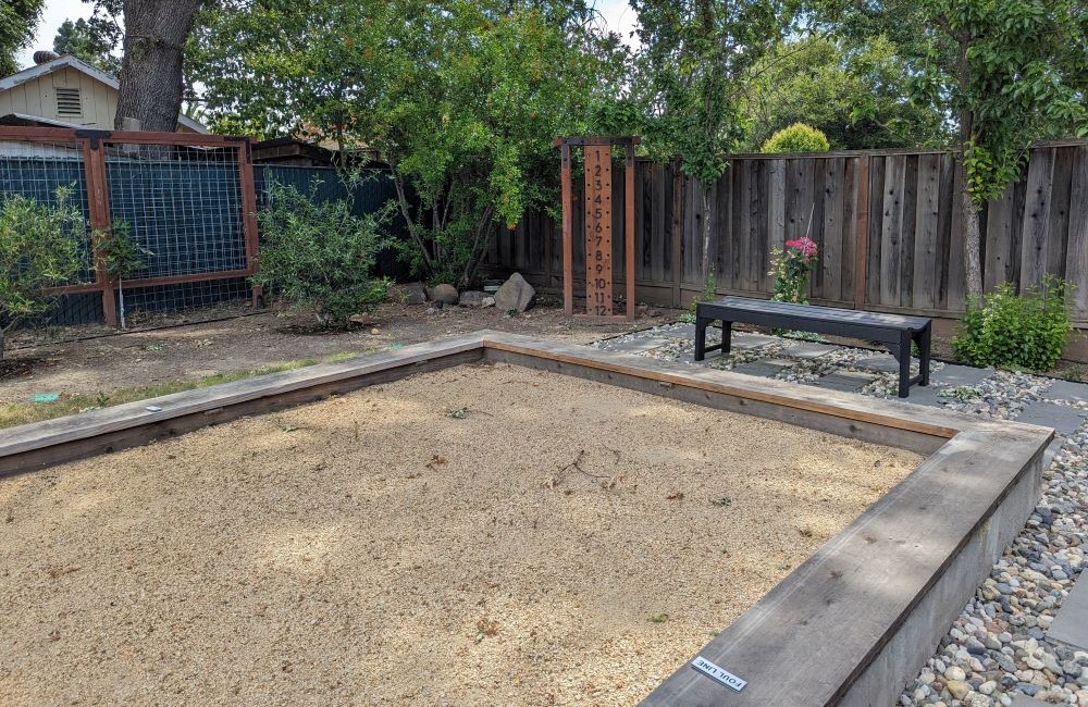 Bocce ball court with riverbed gravel and pavers around. Black bench and scoring tower behind. 