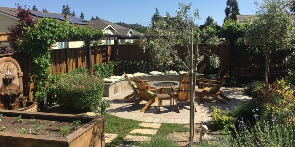 Semicircular stone bench hugs round patio with Adirondack chairs and stone fire pit amid shrubs, trees, groundcover. Grecian fountain in foreground.