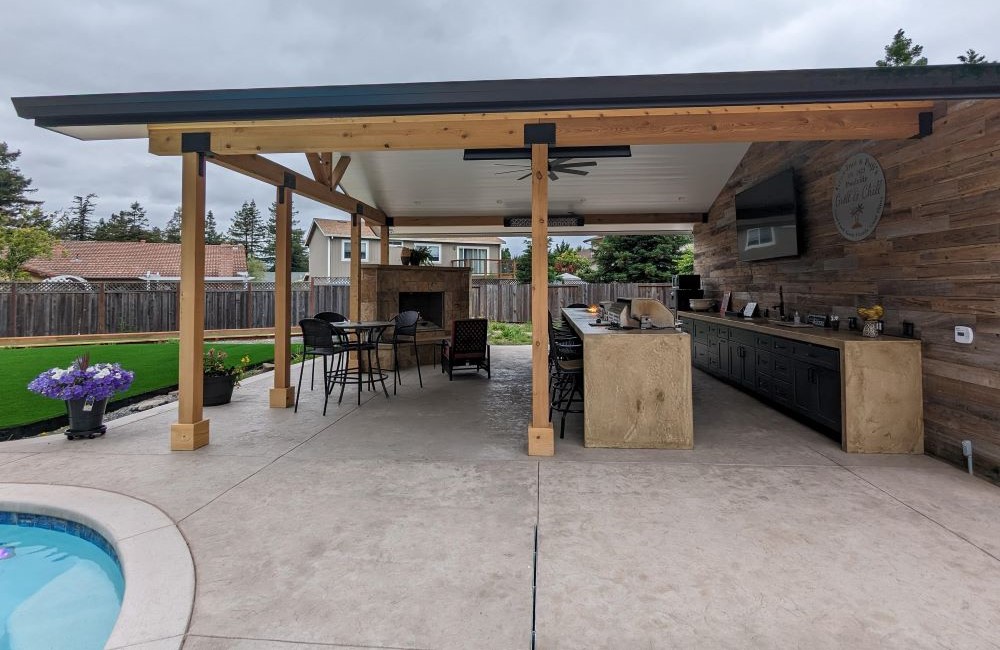 Outdoor covered kitchen with seating area and fireplace shaped fire pit adjacent to lawn and pool. 