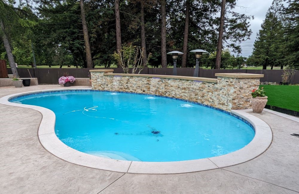 Kidney-shaped pool with rock retaining wall water feature. Tall evergreens in background. 