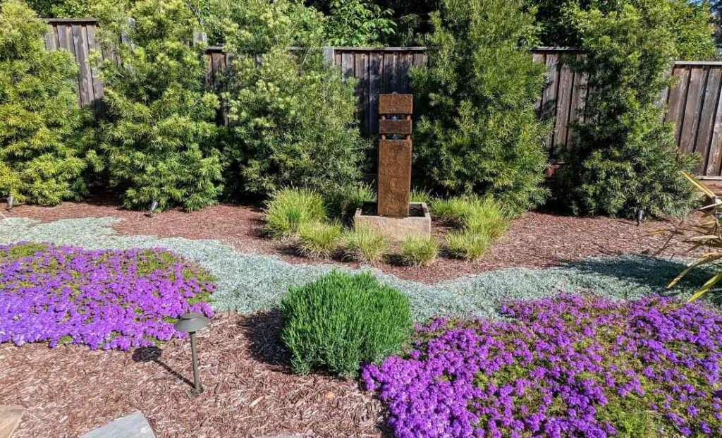 Vertical stone water feature amid native plants. Evergreens line the fence behind and alternating colors of ground cover are in the foreground.